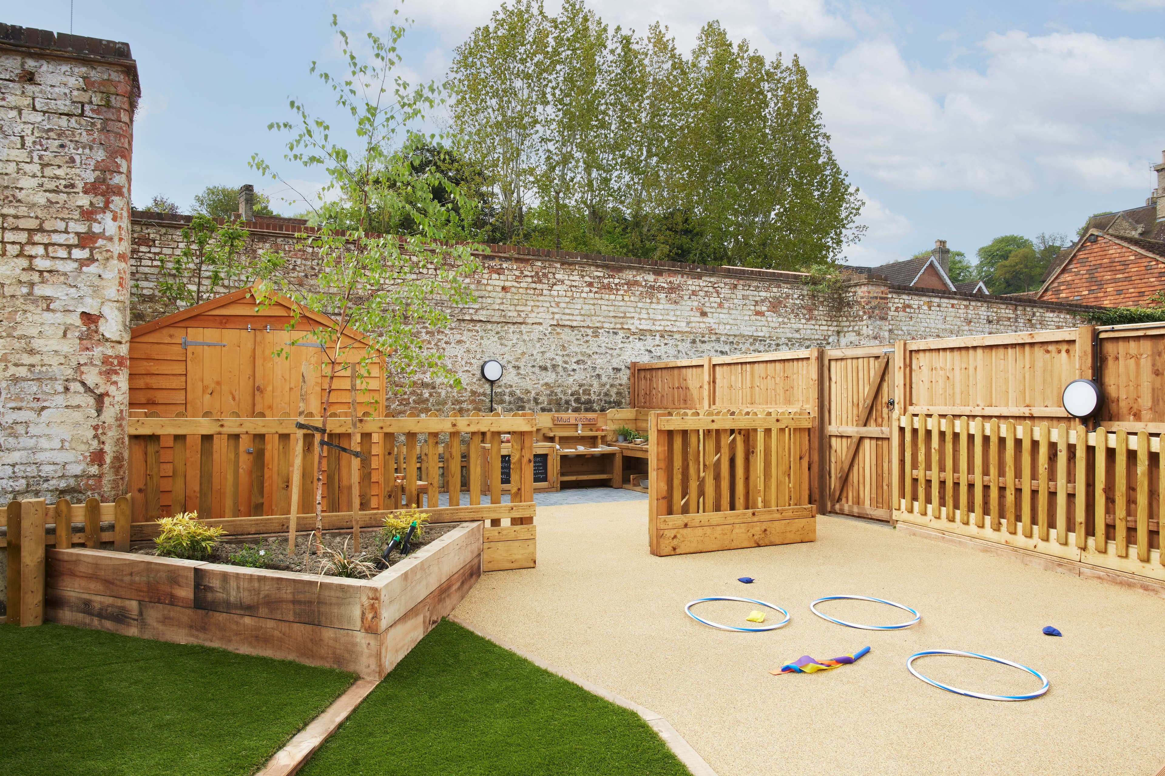 Outdoor play area with a raised garden, wooden shed, fences, and hula hoops on artificial grass and light-colored paving.