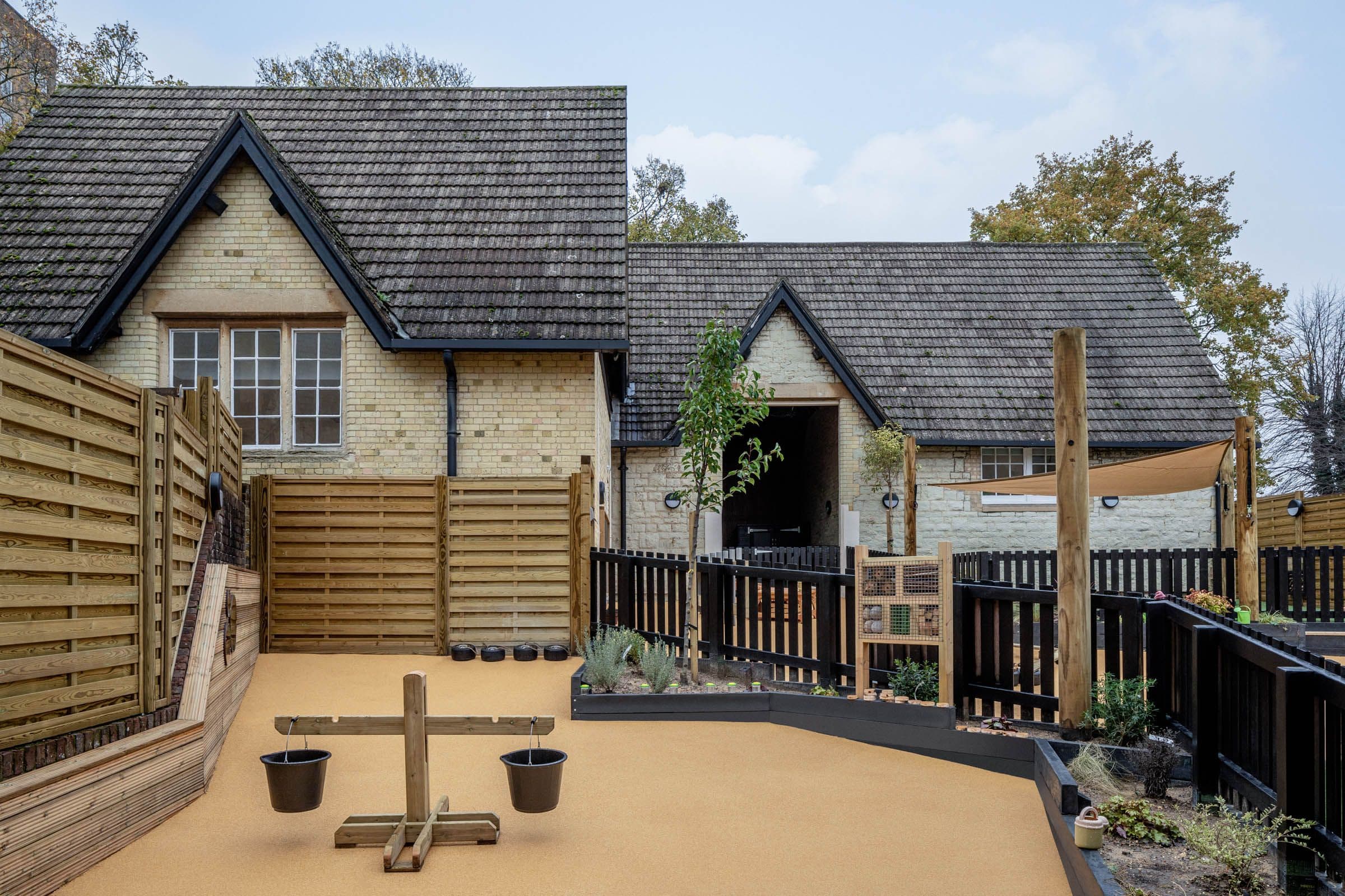 Outdoor play area with a light brick building, soft orange flooring, raised garden beds, and a wooden balance scale.