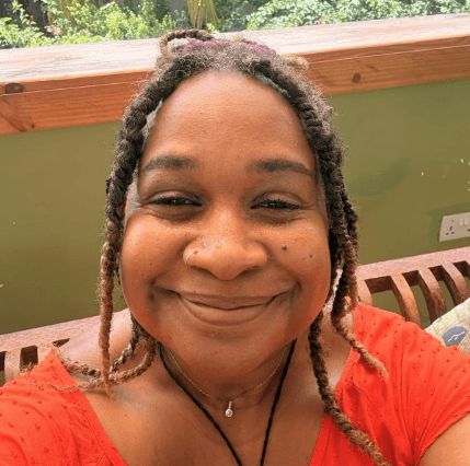 A smiling woman with dreadlocks and a nose ring wears a red top.