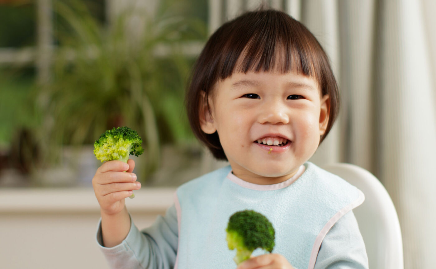 A little girl holding broccoli and smiling